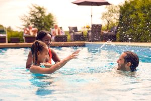 Smiling Mixed Race Family On Summer Holiday Having Fun Splashing In Outdoor Swimming Pool